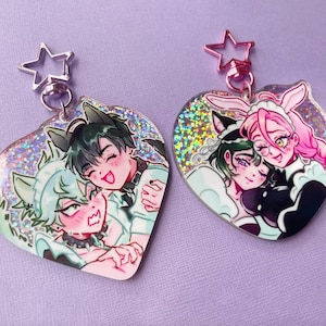 Op de afbeelding: Twee hartvormige sleutelhangers met illustraties in anime-stijl. Eén heeft twee personages met kattenoren, de andere met konijnenoren. Beide hebben een glinsterende, holografische achtergrond en stervormige sluitingen.