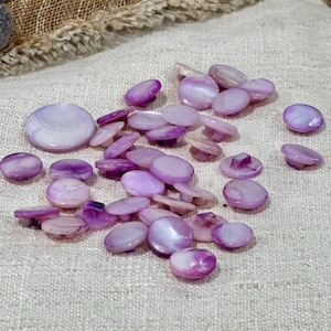 May include: A collection of iridescent purple buttons in various sizes and shapes. The buttons have a pearlescent sheen and are scattered on a neutral-colored fabric. Some buttons have a shank on the back.