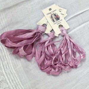 Può includere: Tre fasci di nastro di seta tinto a mano color malva, ciascuno fissato a un cartoncino color crema con l'etichetta Village Curator, un'illustrazione di un ditale e il testo Hand Dyed Silk Ribbon 4mm. I nastri sono drappeggiati in morbidi anelli.