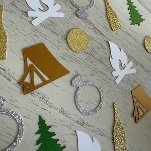 May include: A collection of paper confetti shapes including white campfires, silver glitter engagement rings, gold glitter champagne bottles, green pine trees, gold glitter circles, and gold cardstock tents scattered across a white surface.