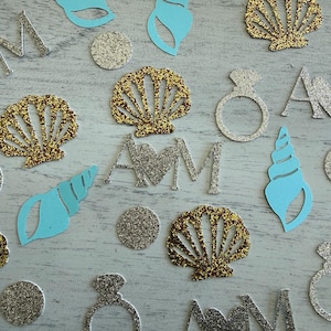 May include: A collection of paper confetti featuring light blue seashell cutouts, gold glitter seashell shapes, silver glitter engagement ring silhouettes, silver glitter circles, and silver glitter letter monograms with a heart in the center.