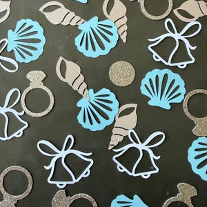 May include: A collection of paper confetti shapes including light blue seashells, tan spiral shells, gold glitter engagement rings, white wedding bells, and small gold glitter circles scattered across the surface for decorative use.