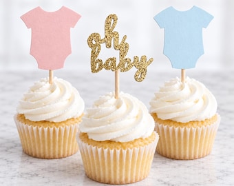 May include: Three cupcakes with white frosting in white paper liners. The left cupcake has a pink baby onesie topper, the right has a light blue baby onesie topper, and the center cupcake has a gold glitter script topper that reads oh baby.