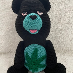May include: A black crocheted teddy bear with a green marijuana leaf on its chest. The bear has a blue face with pink lips and red eyes.