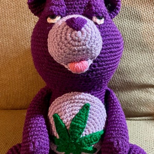 May include: Purple crocheted stuffed bear with a green marijuana leaf on its chest. The bear has a pink tongue sticking out.