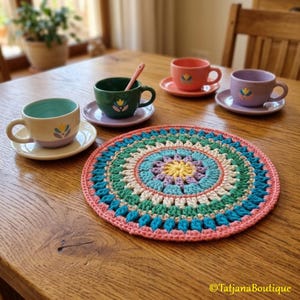 May include: A colorful crochet coaster with a circular design, featuring shades of pink, blue, green, and yellow. The coaster is placed on a wooden table, alongside several teacups and saucers in various colors, including green, orange, and purple.