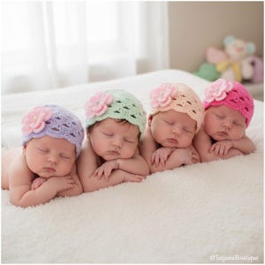 May include: Four newborn babies wearing crocheted hats with flower accents, in shades of lavender, mint green, peach, and hot pink, are lying on a soft, white blanket. The babies are sleeping peacefully, with their arms crossed.