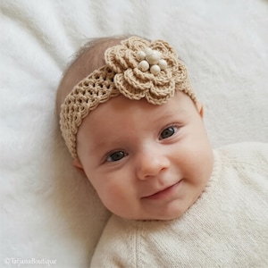 May include: A tan crochet baby headband with a large flower and bead embellishments. The headband is made of a soft, textured yarn and is designed to fit comfortably on a baby's head. The flower is the focal point of the design.