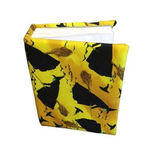 May include: A fabric-covered book featuring a vibrant yellow background with a repeating pattern of black silhouettes of witches on broomsticks, pointed hats, and small bats. The book has white pages visible at the top edge of the binding.