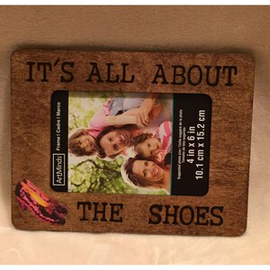 May include: A brown wooden picture frame with the text "IT'S ALL ABOUT THE SHOES" engraved on it. The frame is designed to hold a 4x6 inch photo. A pink shoe is visible in the bottom left corner of the frame.