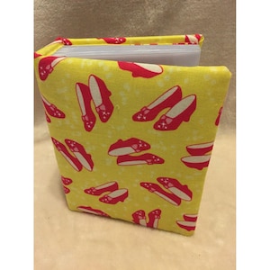 May include: A fabric-covered photo album featuring a bright yellow background with a repeating pattern of red glittery shoes. The album has a visible white plastic spine and internal clear sleeves for holding photographs in a vertical orientation.