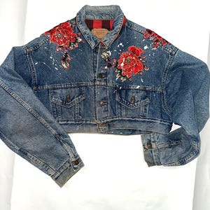 May include: Cropped denim jacket in a medium wash, featuring red floral embroidery on the shoulders and white paint splatters. The jacket has a classic collar, button-up front, and a red and black plaid lining at the collar.