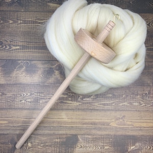 May include: A wooden drop spindle with a brass hook rests on a pile of creamy white wool roving. The spindle has a round wooden whorl and a long, slender shaft. The wool is soft and fluffy, perfect for spinning yarn.