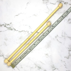 May include: Two light-colored wooden knitting needles with rounded ends are positioned next to a flexible ruler. The needles are of different lengths. The ruler is marked with inch measurements. The background is a white marble pattern.