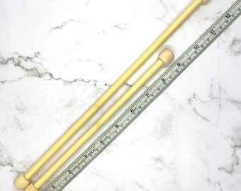 Puede incluir: Dos agujas de tejer de madera de color claro con extremos redondeados se colocan junto a una regla flexible. Las agujas son de diferentes longitudes. La regla está marcada con medidas en centímetros. El fondo es un patrón de mármol blanco.