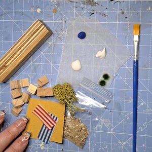May include: A collection of craft supplies including a wooden block, small wooden cubes, paint, moss, and a paintbrush with a blue handle. A small piece of yellow paper with red and blue striped tape is also present. The items are arranged on a blue cutting mat.