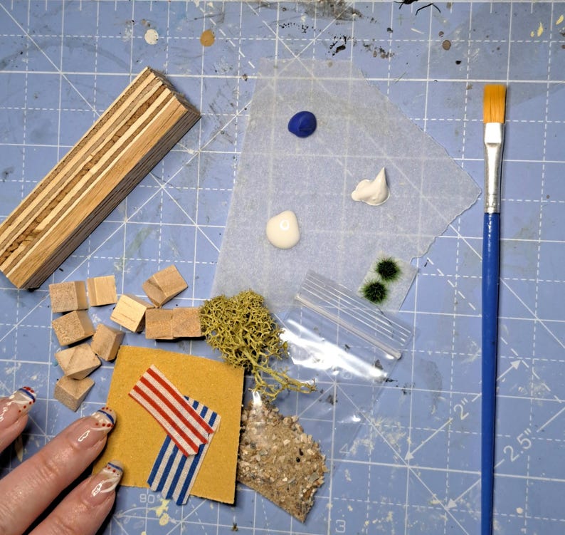 May include: A collection of craft supplies including a wooden block, small wooden cubes, paint, moss, and a paintbrush with a blue handle. A small piece of yellow paper with red and blue striped tape is also present. The items are arranged on a blue cutting mat.