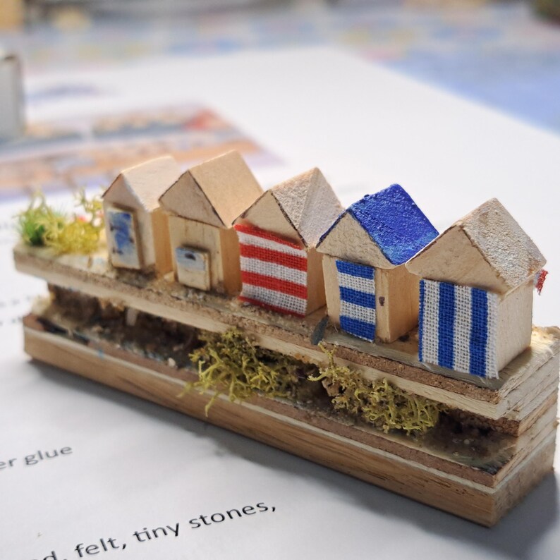 May include: Miniature wooden beach huts with varying colors and patterns. The huts are arranged in a row on a wooden base, with details like red and white stripes, blue doors, and blue and white striped patterns. Green moss adds a natural touch.