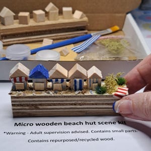 May include: A micro wooden beach hut scene kit. The kit includes miniature wooden beach huts with red, blue, and white accents. The huts are arranged on a wooden base with miniature greenery and a tiny beach towel. The text on the paper reads "Micro wooden beach hut scene kit."