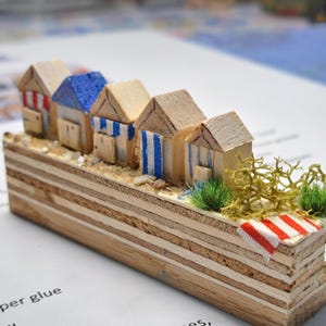 May include: A miniature wooden beach scene featuring five small, colorful beach huts with striped accents. The huts sit on a sandy base with decorative elements, including a miniature red and white striped towel. The base is made of layered wood.