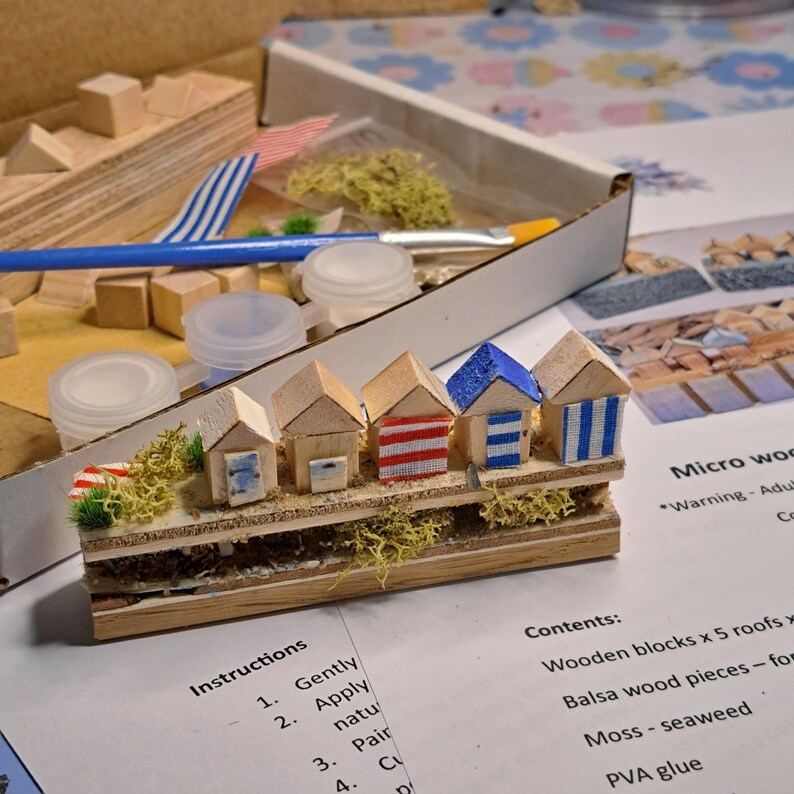 May include: A miniature wooden beach hut kit. The kit includes five small huts with different colored roofs and striped or solid walls. The kit also includes moss, glue, and other craft supplies. The instructions are visible in the image.