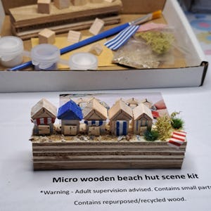 May include: A micro wooden beach hut scene kit. The kit includes miniature wooden beach huts with red, blue, and white accents, set on a wooden base with sand and greenery. The kit also includes paint, brushes, and small wooden blocks.