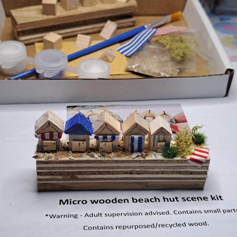 May include: A micro wooden beach hut scene kit. The kit includes miniature wooden beach huts with red, blue, and white accents, set on a wooden base with sand and greenery. The kit also includes paint, brushes, and small wooden blocks.