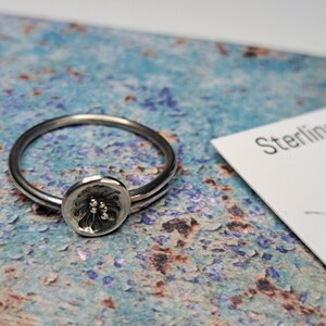 May include: A silver ring with a textured, circular design in the centre. The ring is on a blue and brown textured surface.
