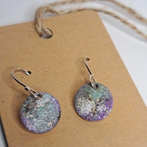 May include: A pair of handmade dangle earrings. Each earring features a circular, textured design with a mix of colors, including purple, teal, and silver. The earrings hang from silver-colored hooks and are displayed on a brown tag.