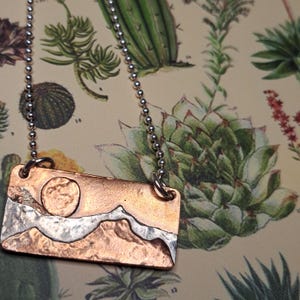 May include: A copper and silver pendant necklace featuring a mountain and sun design. The rectangular pendant hangs from a silver ball chain. The mountains are silver, and the sun is a circle. The background is a copper color.