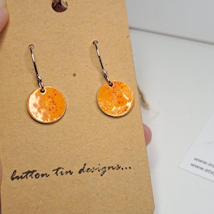 May include: A pair of round, orange earrings with a speckled design. The earrings are attached to silver-colored hooks. The earrings are displayed on a brown card with the text "button tin designs..." written in cursive.