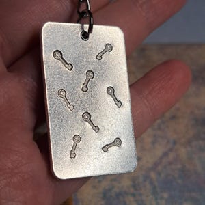 May include: A rectangular silver metal pendant with a series of small key-shaped designs etched into its surface. The pendant is attached to a metal chain. The key designs are evenly spaced across the pendant.