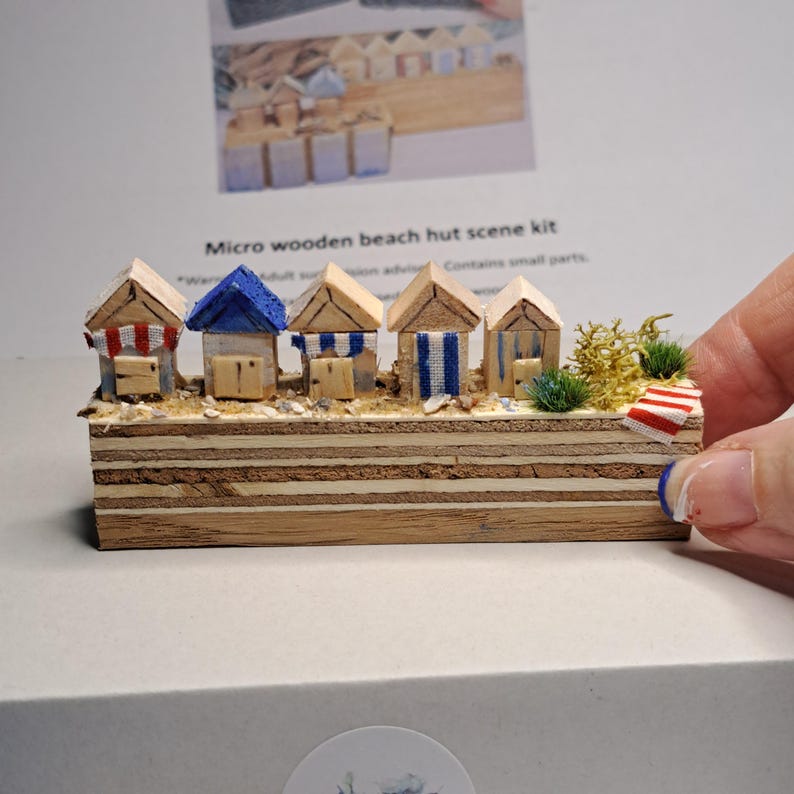 May include: A miniature wooden beach hut scene kit. The scene features five small huts with varying roof colors and striped awnings. The huts are set on a sandy base with small plants and a miniature beach towel. The text "Micro wooden beach hut scene kit" is visible.