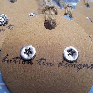 May include: Close-up of two silver-colored star-shaped stud earrings on a brown tag. The earrings are round with a star design in the center. The tag has the words "button tin designs" written on it.