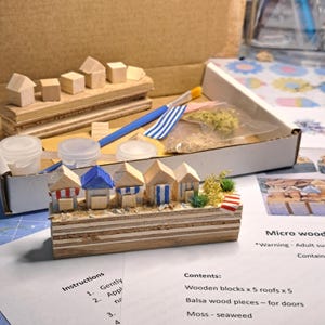 May include: A miniature beach hut kit with wooden blocks, roofs, and balsa wood pieces. The kit includes a paintbrush, paint pots, and moss. The huts have blue, red, and white accents. The kit is designed for adult supervision.