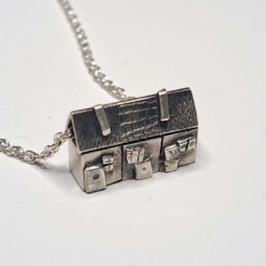 May include: A silver-colored house pendant necklace. The pendant is a miniature house with detailed features, including a textured roof, windows, and doors. The necklace chain is also silver.