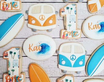 May include: A set of decorated sugar cookies featuring a beach theme. The cookies are decorated with images of surfboards, palm trees, and vintage vans. The cookies are also decorated with the words "Kai" and "One".