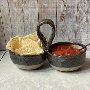 May include: A ceramic chip and dip serving dish with a handle. One side holds tortilla chips, the other salsa. The dish is dark brown with a speckled design and a light tan base. A black spoon rests in the salsa.