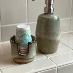 A Square Style Cup Dispenser and a Barrel Style Soap Pump, both in Seafoam Green, sit on a bathtub or sink edge, ready for use.