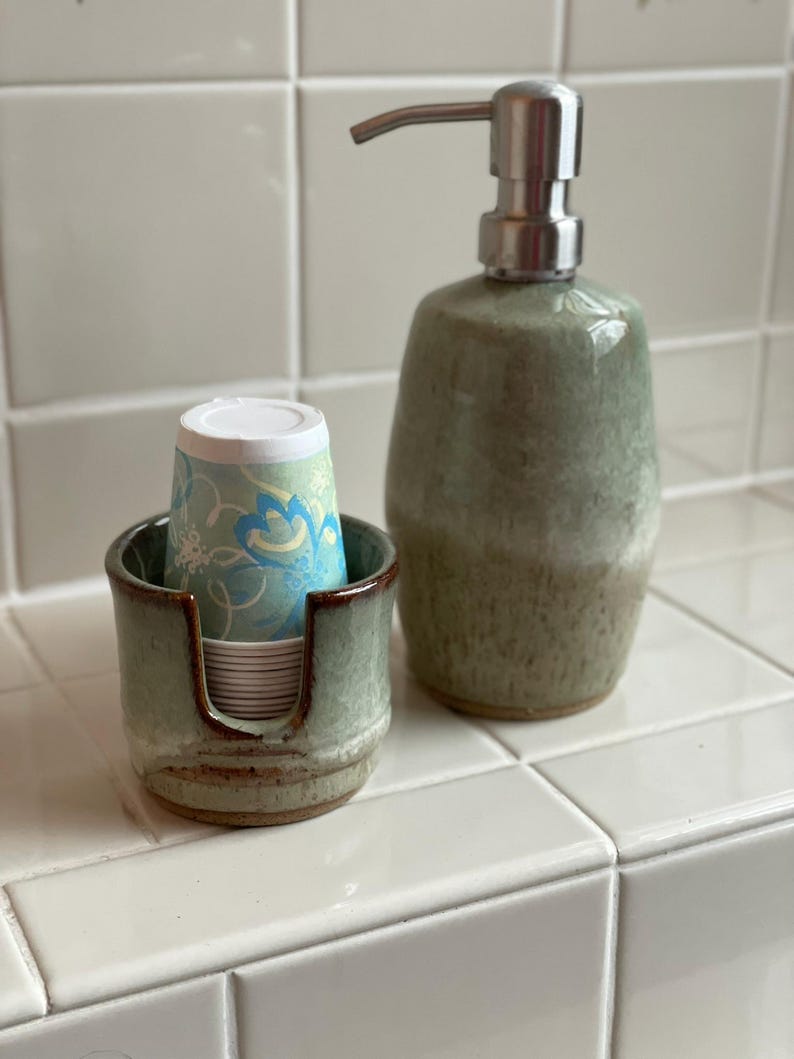 A Square Style Cup Dispenser and a Barrel Style Soap Pump, both in Seafoam Green, sit on a bathtub or sink edge, ready for use.