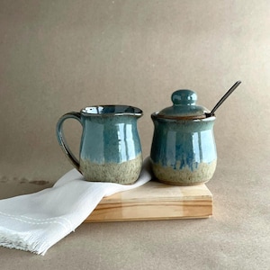 May include: A teal and tan ceramic creamer and sugar bowl set with a lid and spoon. The set is on a wooden tray with a white cloth. The creamer has a handle and the sugar bowl has a rounded shape.