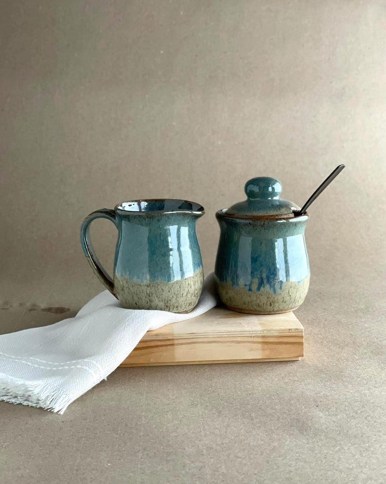 May include: A teal and tan ceramic creamer and sugar bowl set with a lid and spoon. The set is on a wooden tray with a white cloth. The creamer has a handle and the sugar bowl has a rounded shape.