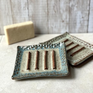 May include: Two ceramic soap dishes with a rectangular shape and a light blue and beige color scheme. Each dish has four brown bars and a textured border. A bar of soap is visible in the background.