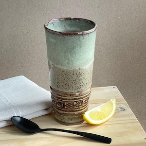 Könnte beinhalten: Ein handgefertigter Keramikbecher mit zweifarbigem Grün- und Braunglasur, mit einem dekorativen Band am Boden. Ein schwarzer Löffel, eine Zitronenscheibe und ein gefaltetes weißes Tuch liegen auf einem Holztablett.