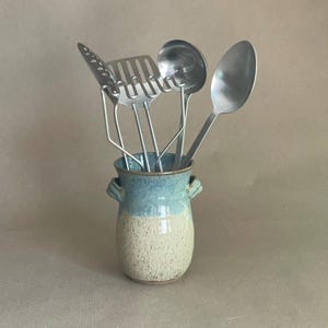 Puede incluir: Un recipiente de cerámica para utensilios con un esmalte azul claro y beige, que contiene varios utensilios de cocina de acero inoxidable. Las herramientas incluyen una espátula ranurada, un cucharón y una cuchara. El recipiente tiene dos asas pequeñas.