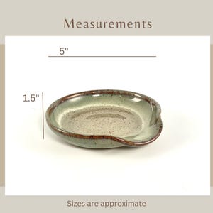 Titled Measurements. A ceramic spoon rest with a speckled gray interior and a green and brown rim. The spoon rest is approximately 5 inches long and 1.5 inches wide. Words on the bottom says sizes are approximate.