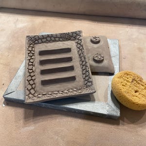 Two in process handmade clay soap dishes. The top one is a rectangular piece with four horizontal slots. The bottom one is up side down showing two of four feet. And a yellow sponge. The unglazed pottery is a light brown color.