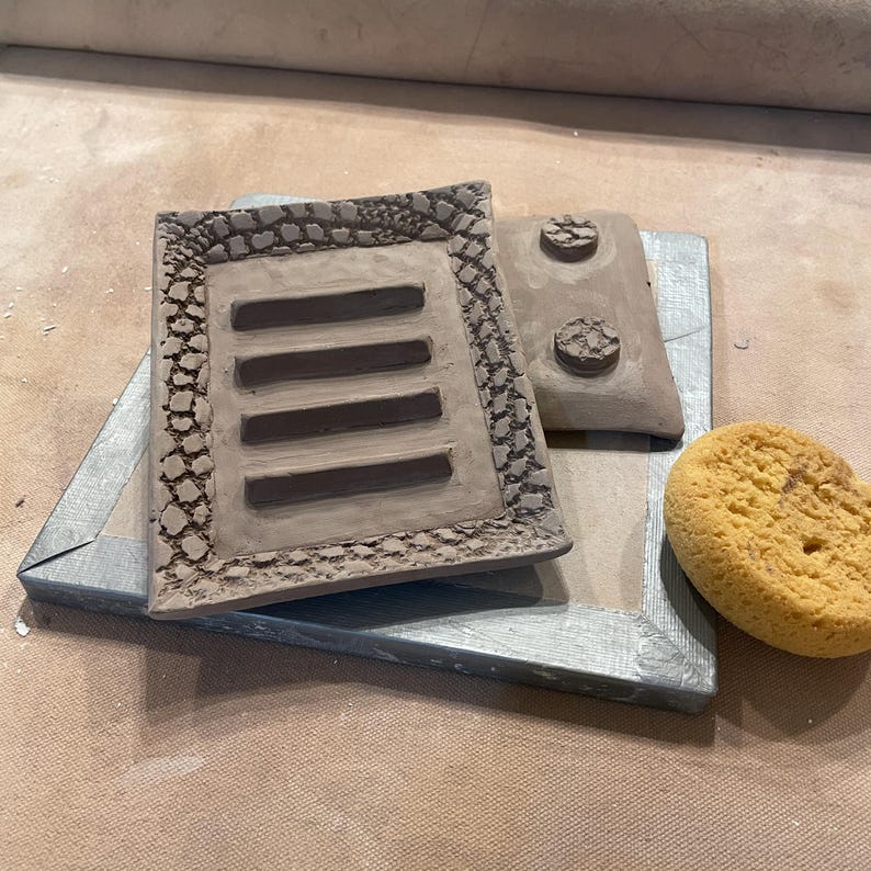 Two in process handmade clay soap dishes. The top one is a rectangular piece with four horizontal slots. The bottom one is up side down showing two of four feet. And a yellow sponge. The unglazed pottery is a light brown color.