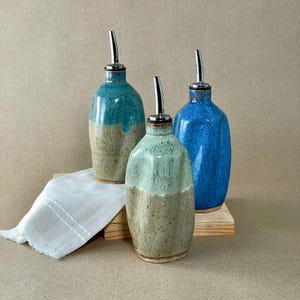 May include: Three ceramic oil dispensers with metallic pour spouts. One is teal and beige, one is light green and beige, and one is solid blue. Each bottle has a rounded shape and a speckled glaze finish. A white cloth is folded underneath.