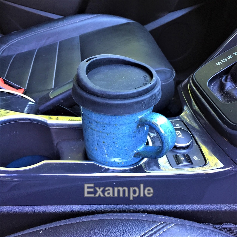 May include: A blue ceramic mug with a black lid sits in a car's cup holder. The mug has a handle and a speckled texture. The word "Example" is printed on the car's console. The interior of the car is visible.